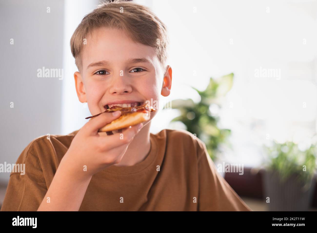 Zufriedener Junge genießt es, leckere Pizza in der Küche zu Hause zu essen. Stockfoto