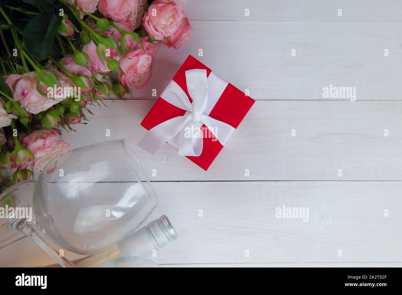 Ein großer Strauß rosafarbener Rosen, ein Geschenk in einer roten Schachtel und eine Flasche Rosenwein mit Gläsern liegen auf der Seite auf einem weißen Holzhintergrund mit einem Platz für Text Stockfoto