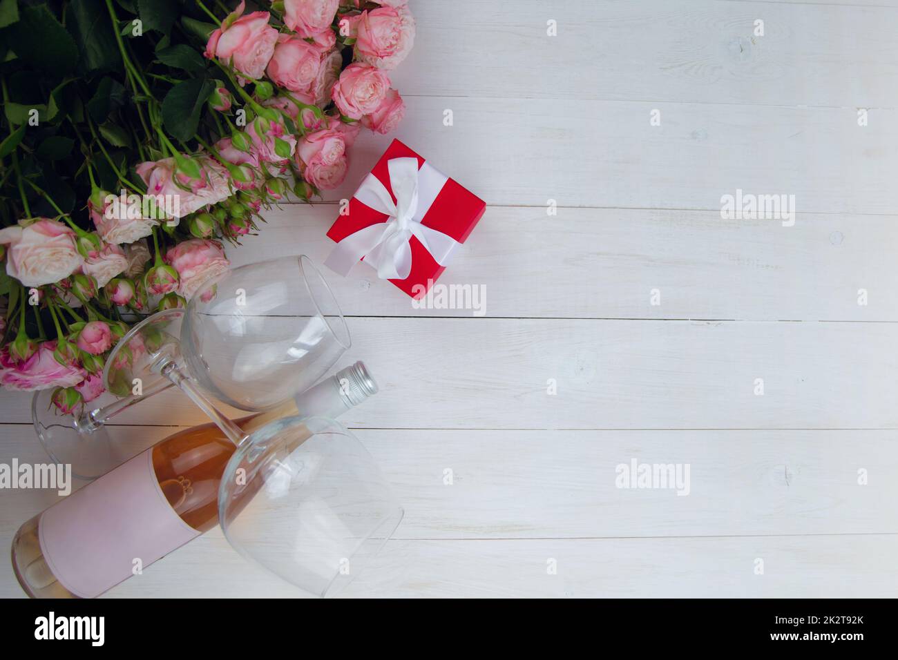 Ein großer Strauß rosafarbener Rosen, ein Geschenk in einer roten Schachtel und eine Flasche Rosenwein mit Gläsern liegen auf der Seite auf einem weißen Holzhintergrund mit einem Platz für Text Stockfoto