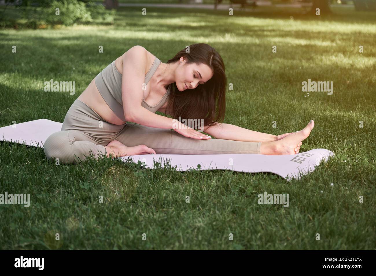 Eine junge Frau übt an einem warmen Sommertag Yoga in einem Stadtpark. Sportlerin sitzt auf einer Turnmatte. Stockfoto