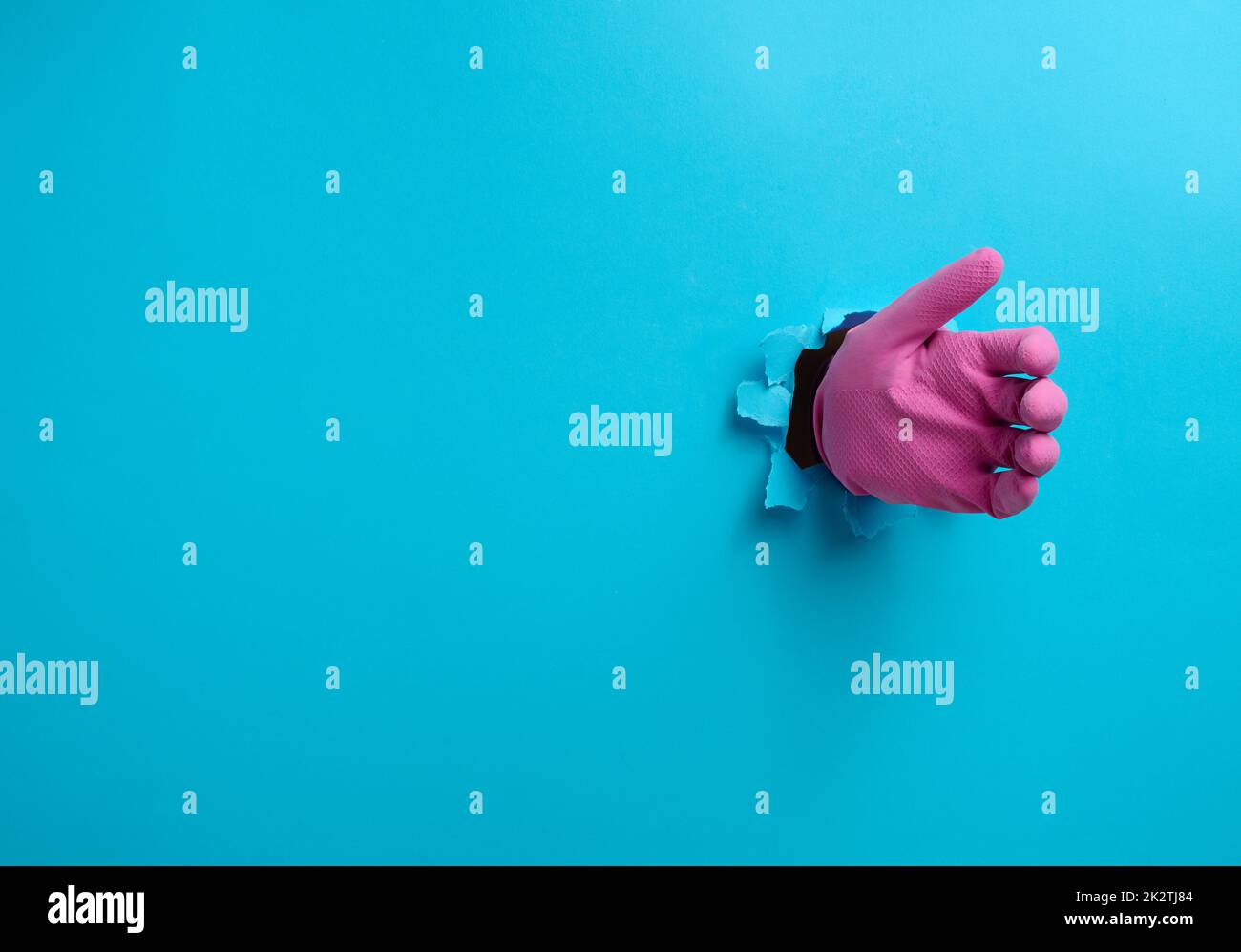 Eine Hand in einem pinken Latexhandschuh hält ein Objekt, ein Teil des Körpers ragt aus einem zerrissenen Loch auf einem blauen Papierhintergrund heraus Stockfoto