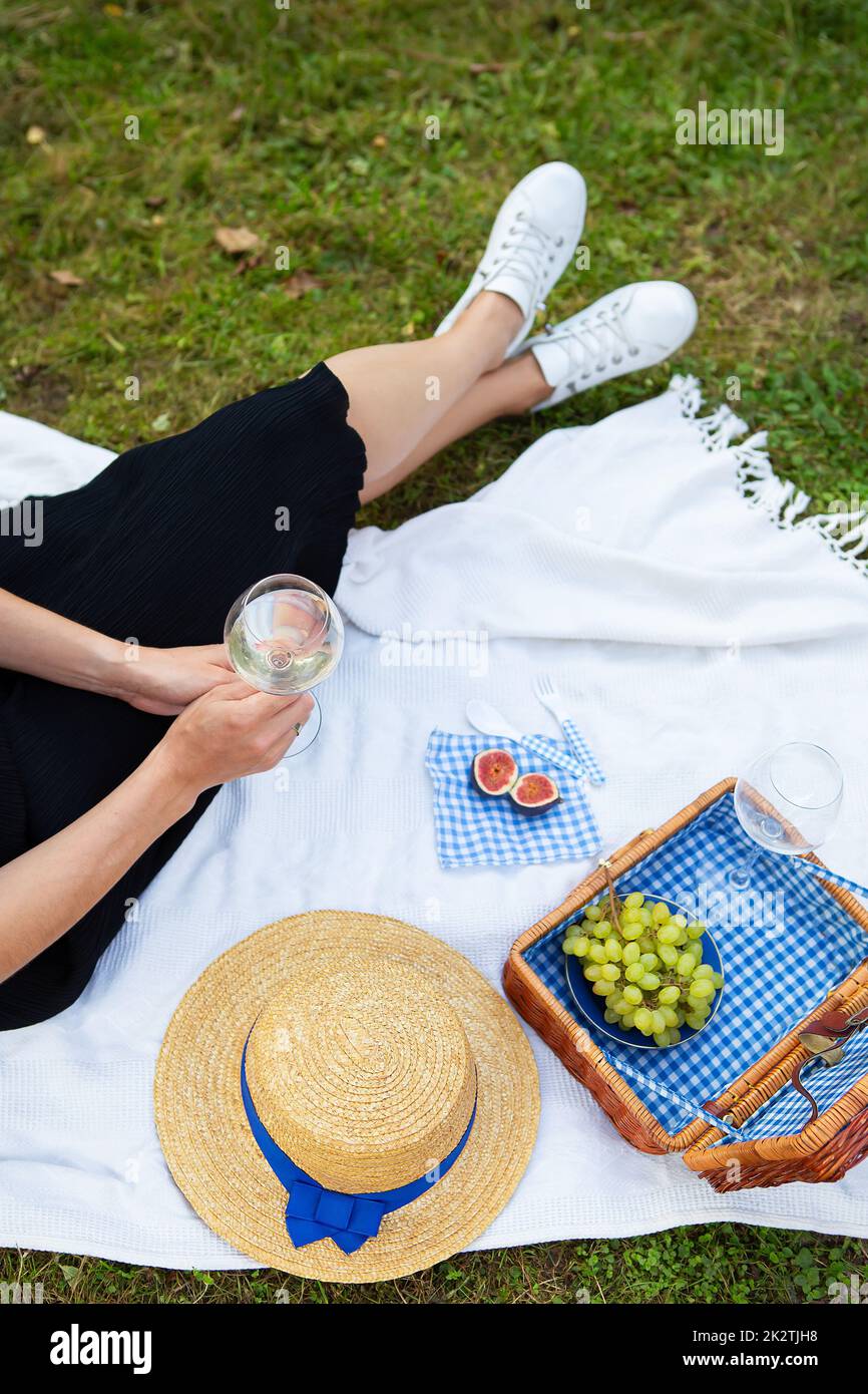 Romantisches Picknick im Park auf dem Gras, köstliches Essen: Korb, Wein, Trauben, Feigen, Käse, blaue karierte Tischdecke, zwei Gläser Wein. Das Mädchen hält ein Glas Wein in den Händen. Das Konzept der Outdoor-Erholung. Stockfoto