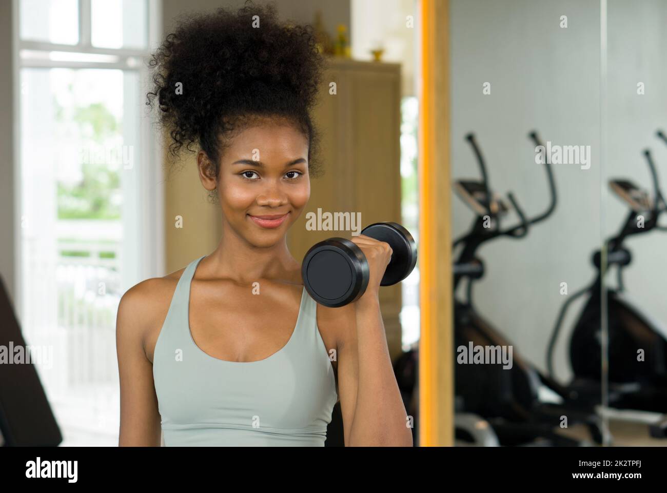 Junge Frau mit lockigen schwarzen Haaren genießt ihren Urlaub im Fitnesscenter. Hebe die Langhantel mit einer Hand und lächle dabei. Stockfoto