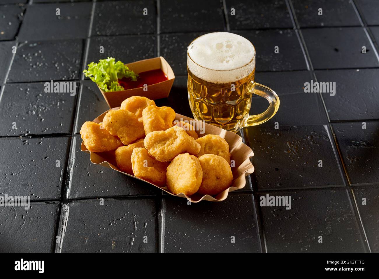 Ein Glas Bier neben dem Teller mit Chicken Nuggets auf dem schwarzen Tisch Stockfoto