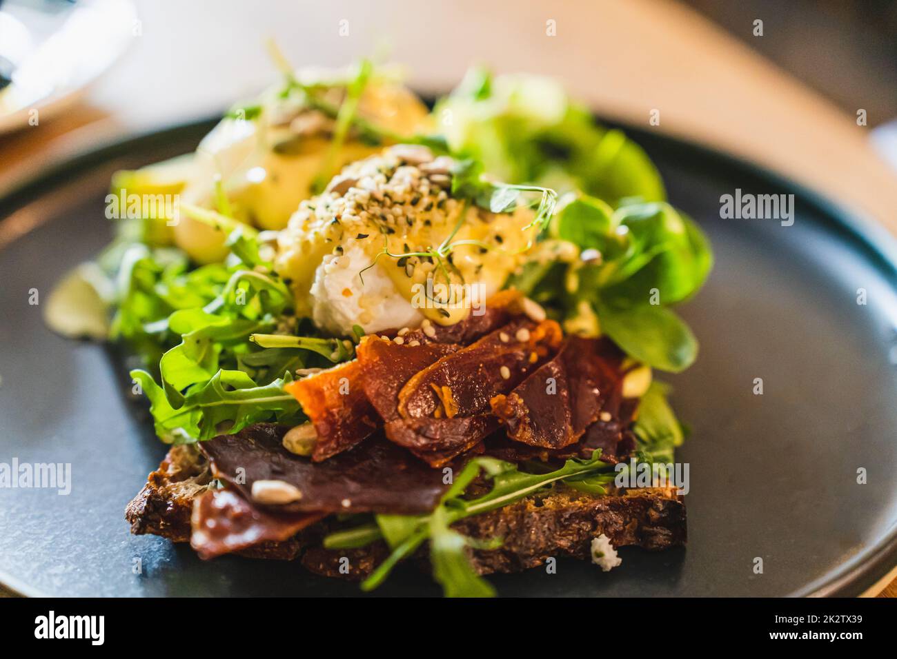 Eier benedict mit getrocknetem Schinken und Salatmischung auf einem schwarzen Teller. Nahaufnahme, selektiver Fokus. Stockfoto