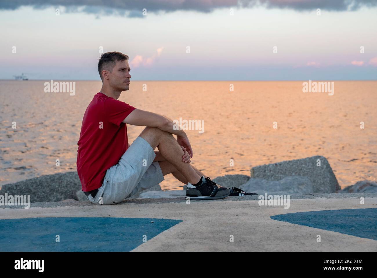 Nachdenklicher junger Mann, der sich an der Promenade mit dem Strand im Hintergrund ausruhte Stockfoto