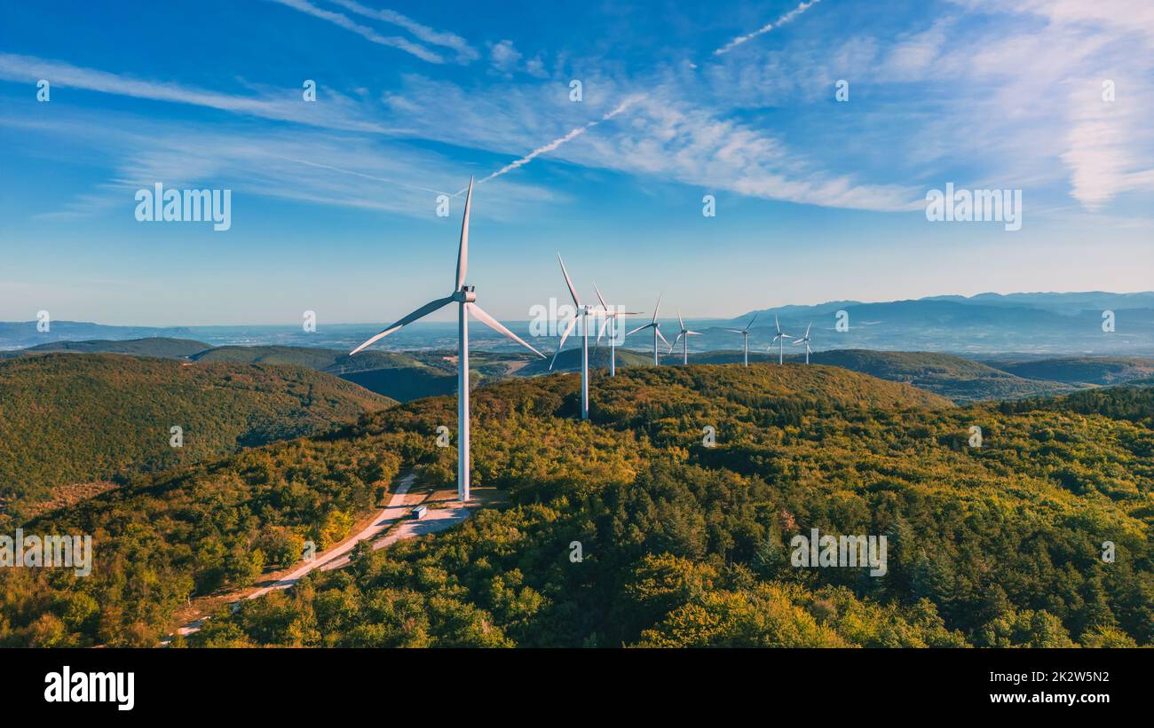 Blick auf einen Windpark in einem bergigen Waldfeld mit Bergen im Hintergrund. Blick während der aufgehenden Sonne. Sonnenaufgang. Stockfoto