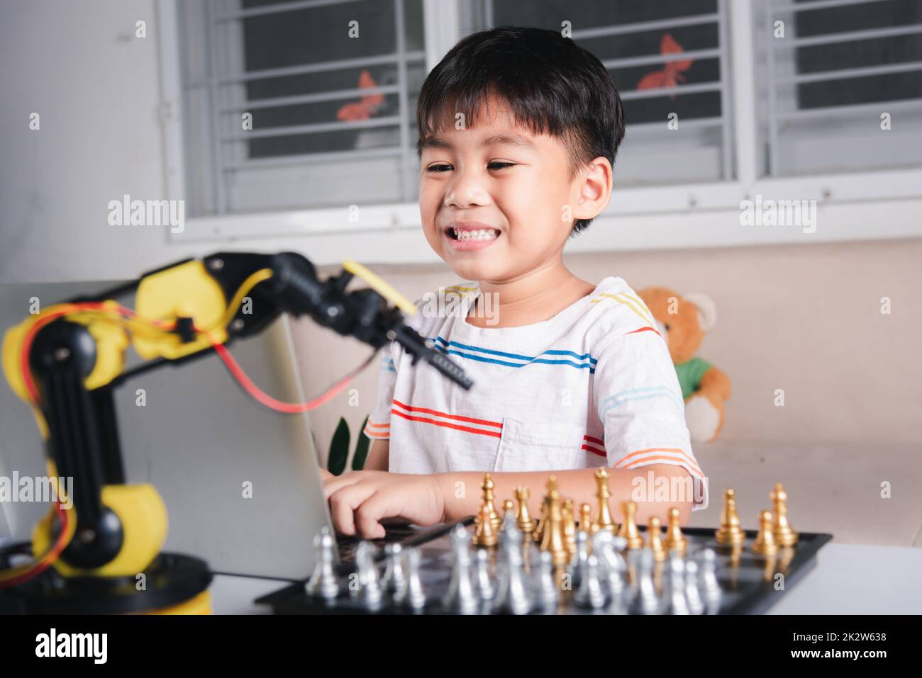 Asiatischer kleiner Junge programmiert Code für Robotermaschinenarm auf Laptop für Schachspiel Stockfoto