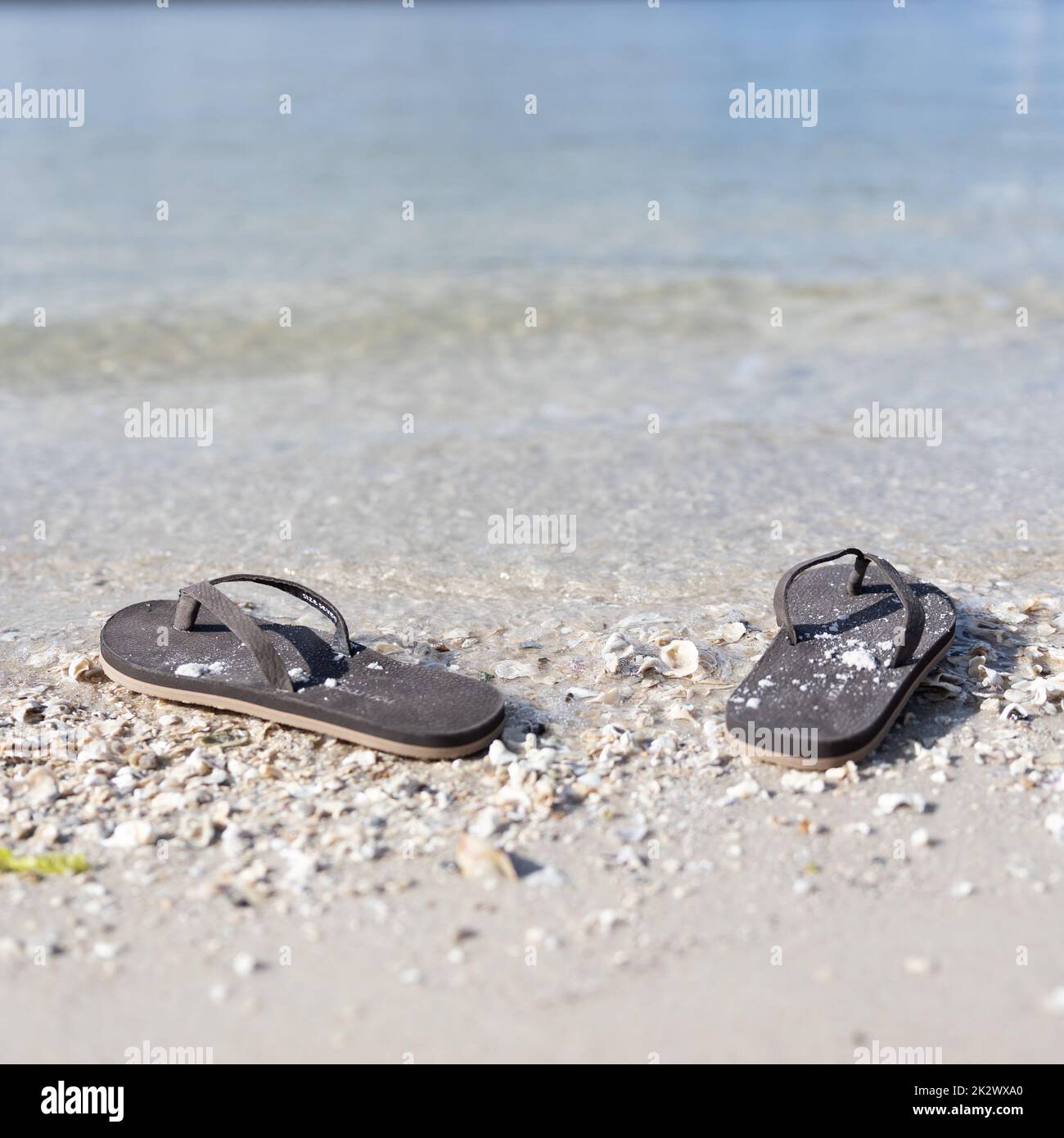 Ein Paar schwarze Flipflops mit weißem Sand am Strand, eine sanfte Flut, die sich hinter ihnen hin und her bewegt Stockfoto