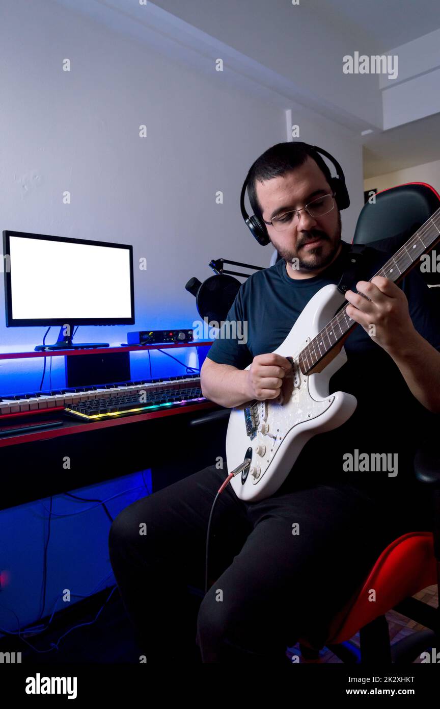 Bärtiger, männlicher Rockmusikproduzent mit Brille und Kopfhörer, der die E-Gitarre in seinem Heimmusikstudio aufnahm. Stockfoto