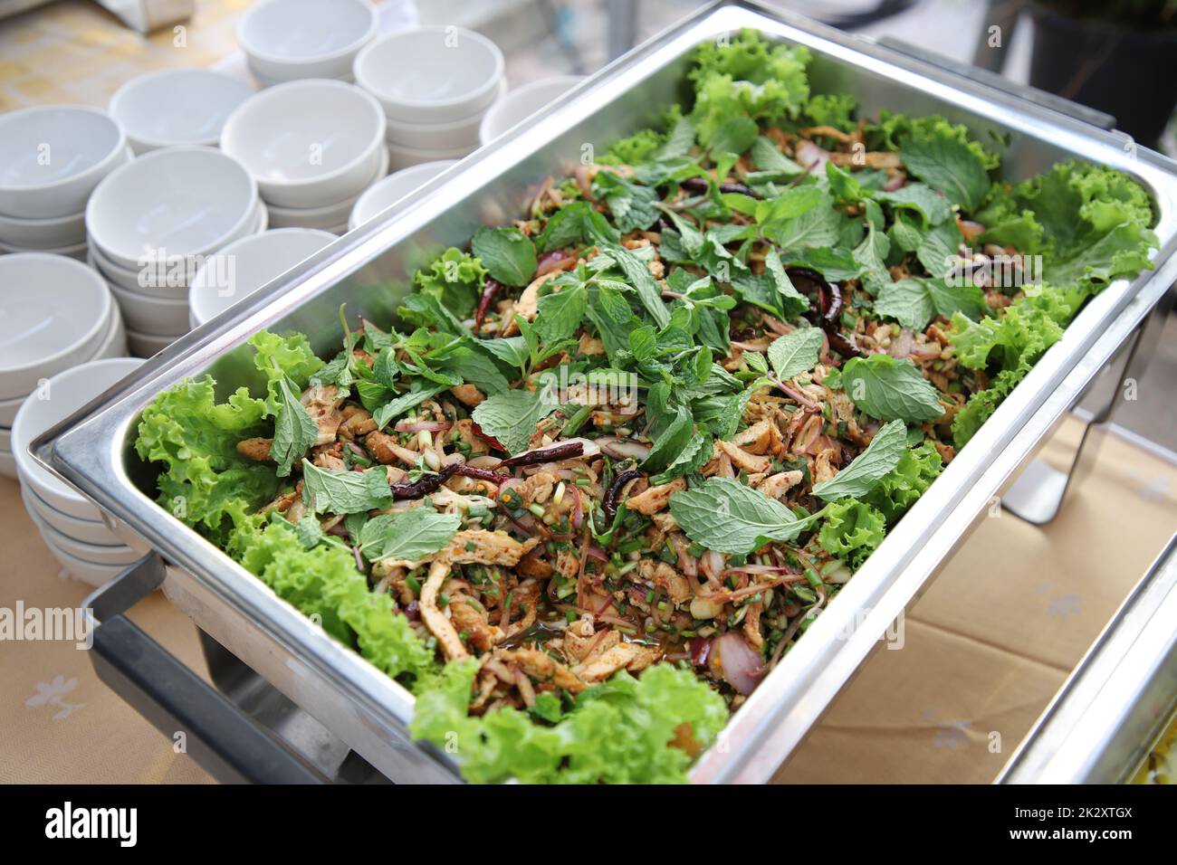 Larb Moo, oben mit Minzeblättern in Buffetschlange bestreut. Der Behälter ist ein rechteckiges Edelstahlfach mit kleinen weißen Tassen, die an der Seite angeordnet sind. Selektiver Fokus. Stockfoto
