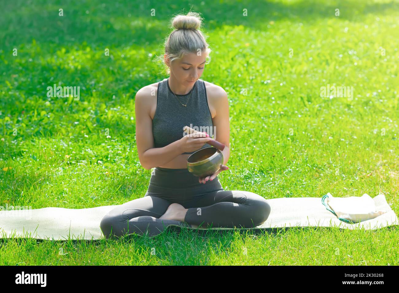 Eine schlanke Frau sitzt auf dem grünen Gras, hält eine Meditationsschale, treibt einen Stock darauf. Stockfoto