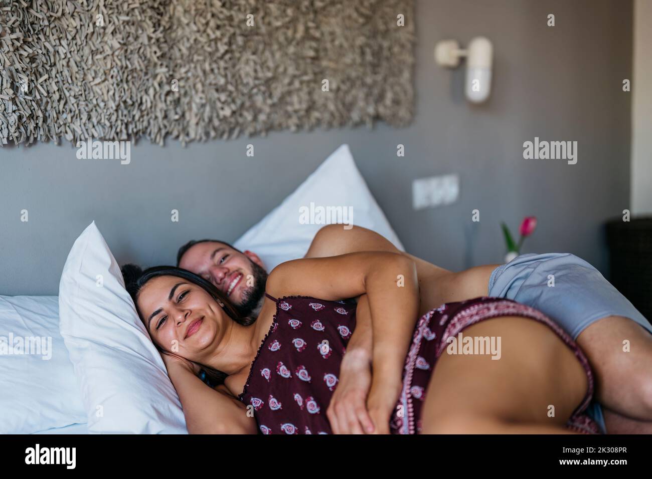 Lächelnde lateinische Braut und Bräutigam blicken auf die Kamera, während sie auf einem Bett liegen Stockfoto