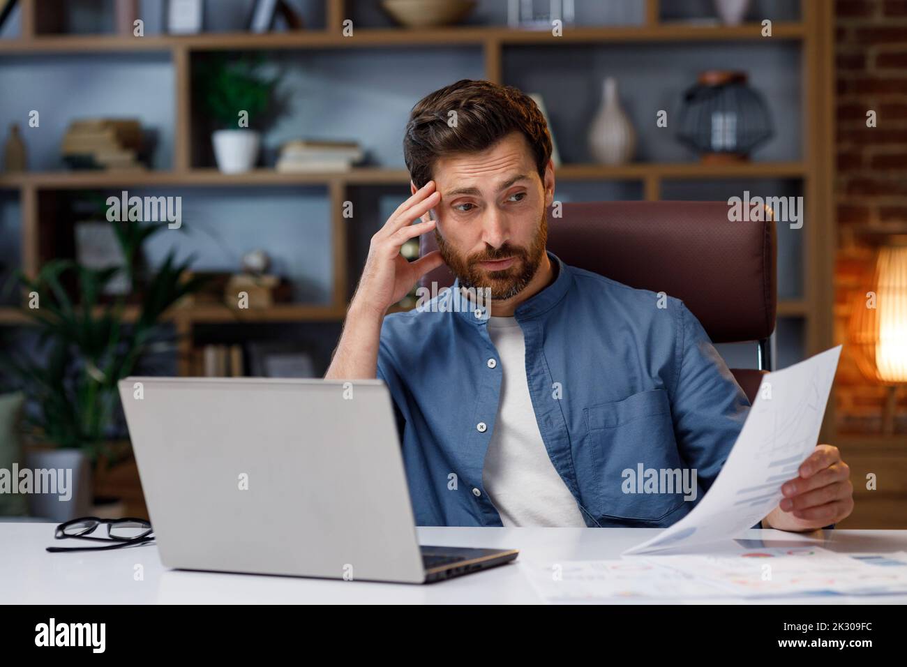 Schöner Mann, der am Laptop arbeitet, während er im Heimbüro sitzt. Gestresst betrachtet er die Verkaufscharts, die Finanzkrise an der Börse. Schlecht Stockfoto