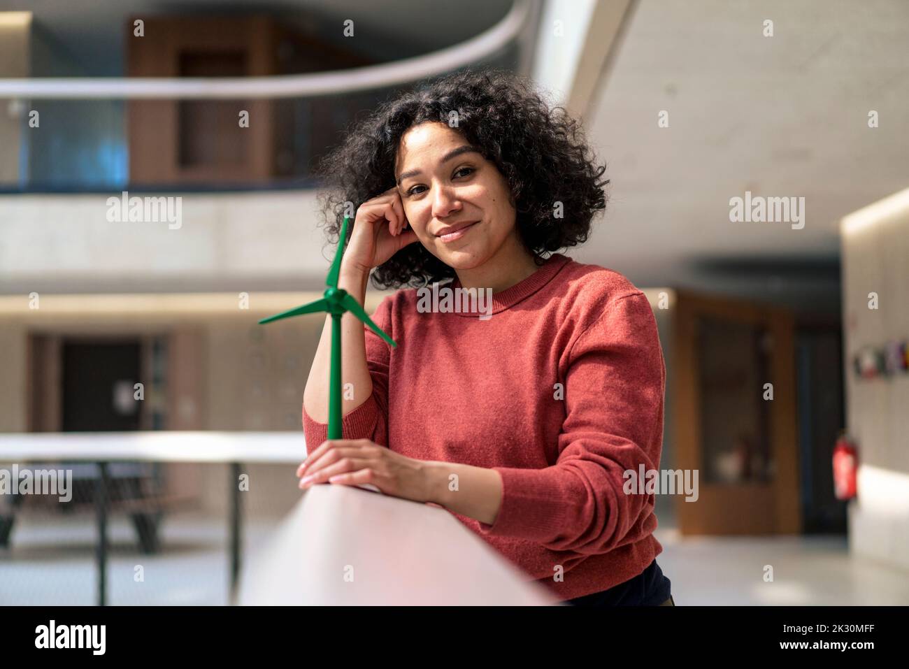 Lächelnde Geschäftsfrau mit Kopf in der Hand, die die Windenergieanlage auf dem Geländer im Büroflur hält Stockfoto