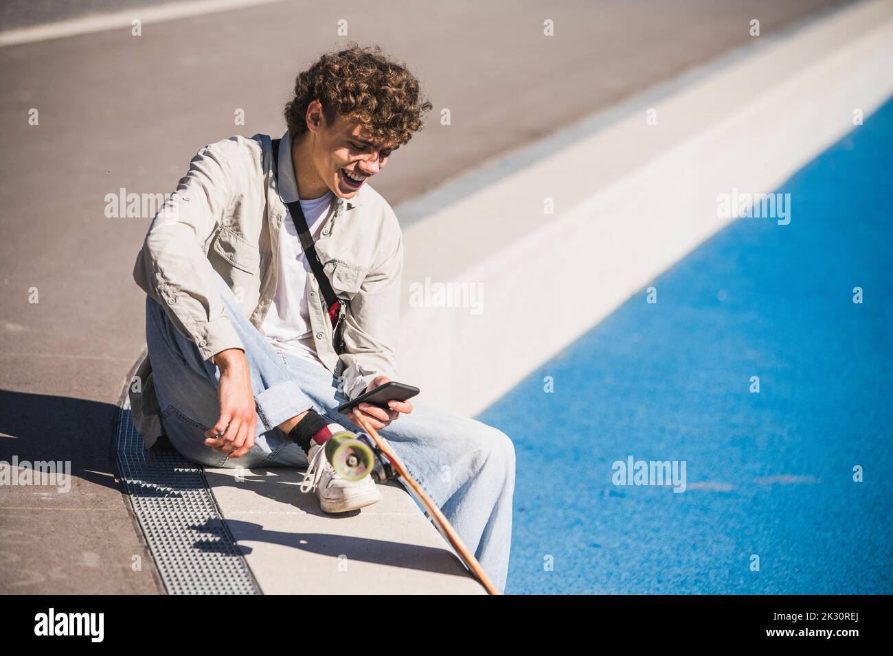 Glücklicher Mann mit Skateboard mit Smartphone am Rand von Strecke und Feld Stockfoto