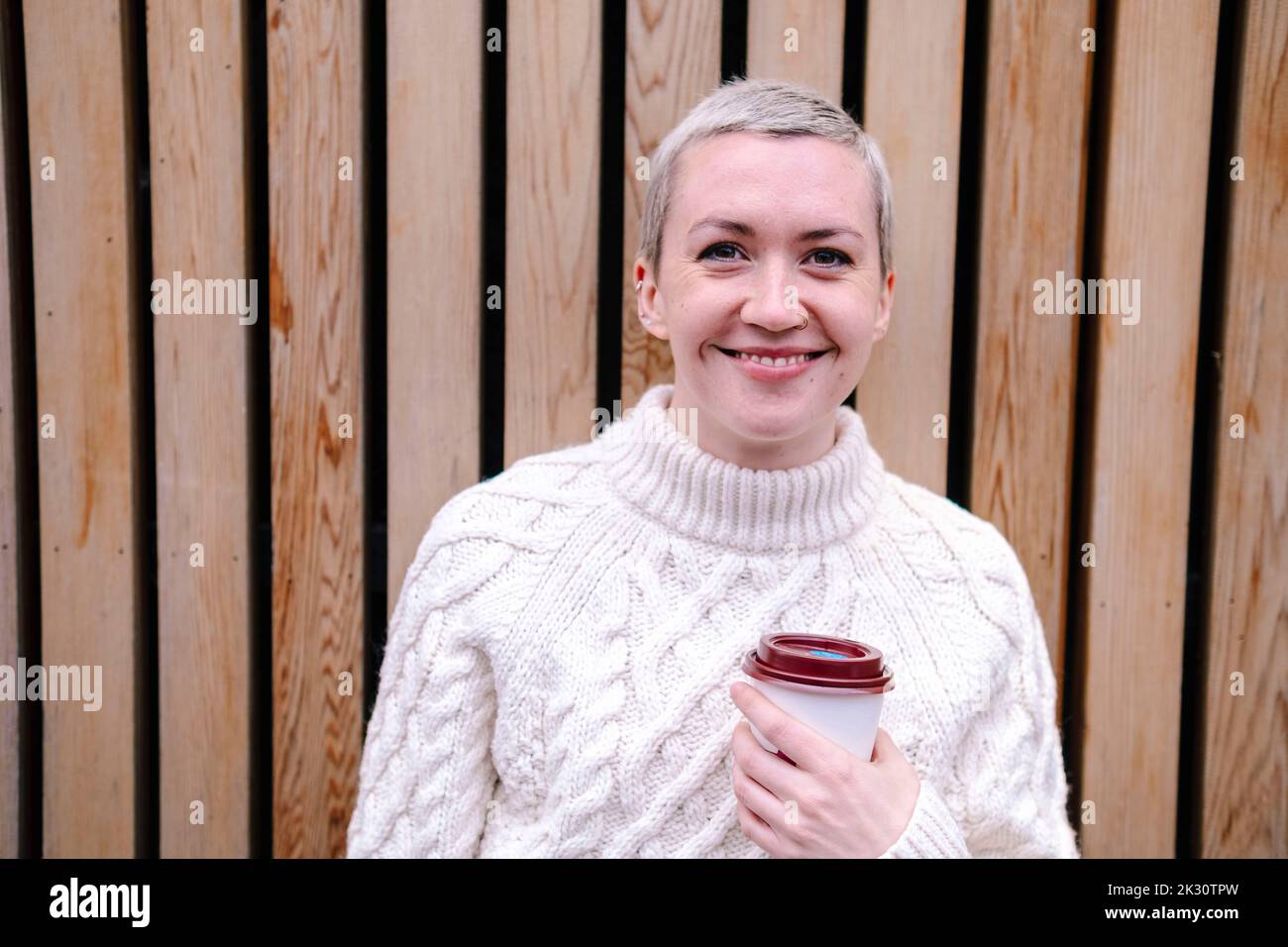 Lächelnde Frau mit kurzen Haaren, die den wiederverwendbaren Becher vor der Holzwand hält Stockfoto