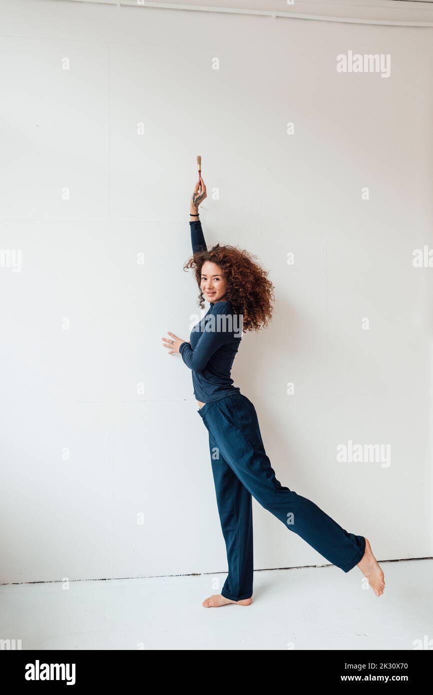 Junge Frau mit erhobener Hand, die den Pinsel auf einem Bein vor der weißen Wand hält Stockfoto