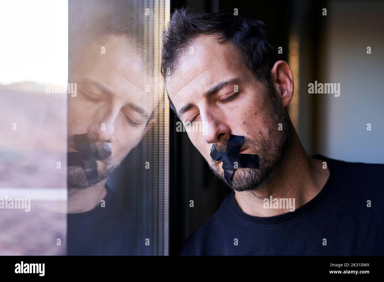 Mann mit Klebeband in Kreuzform, der den Mund auf das Fenster stützt Stockfoto