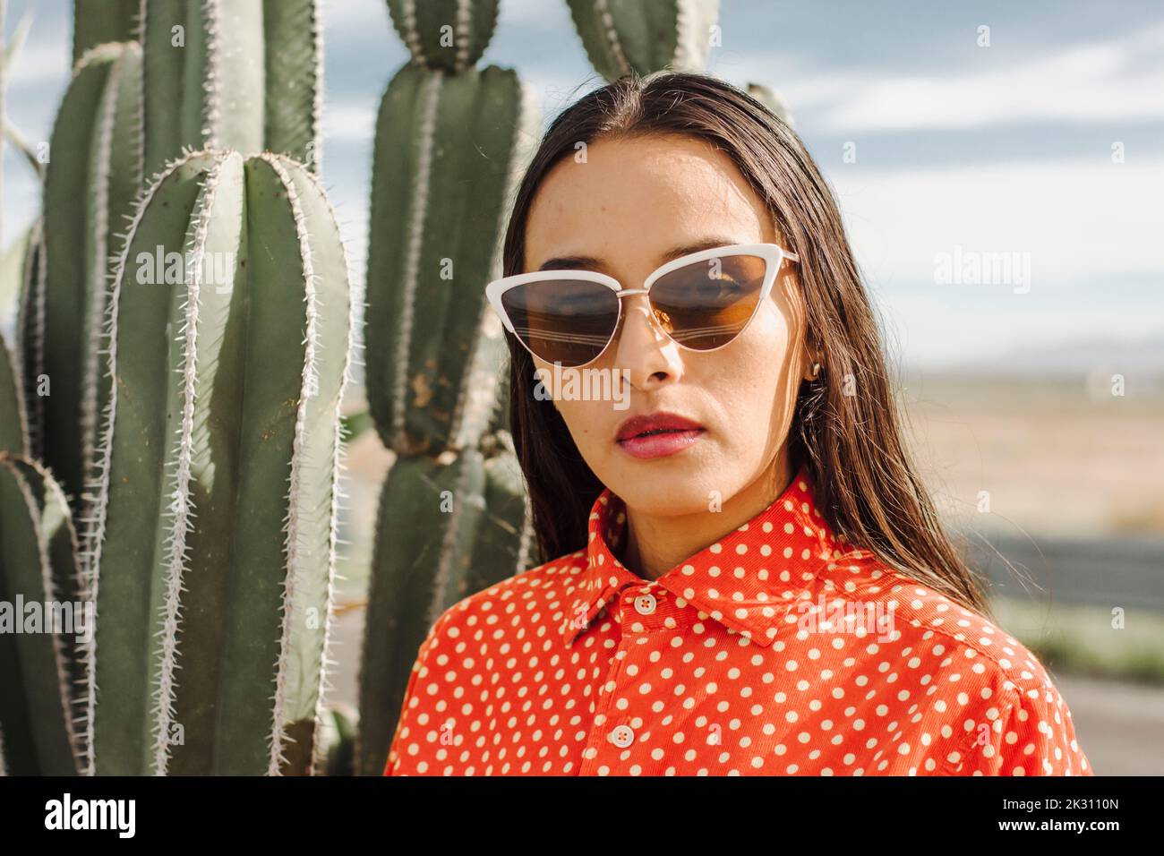 Selbstbewusste junge Frau mit Sonnenbrille, die vor einer Kaktuspflanze steht Stockfoto