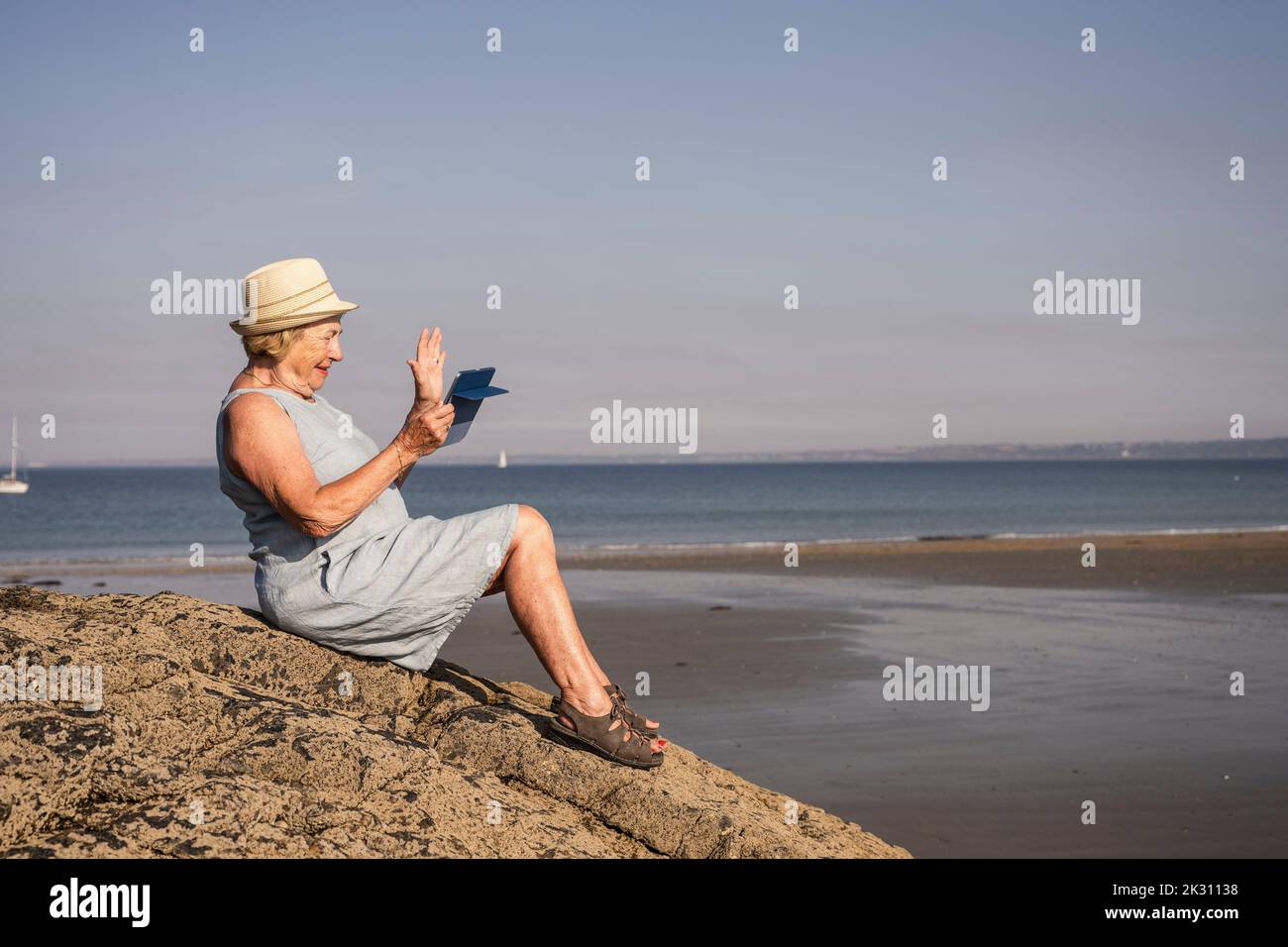 Lächelnde ältere Frau, die bei einem Videoanruf am Strand mit der Hand winkt Stockfoto