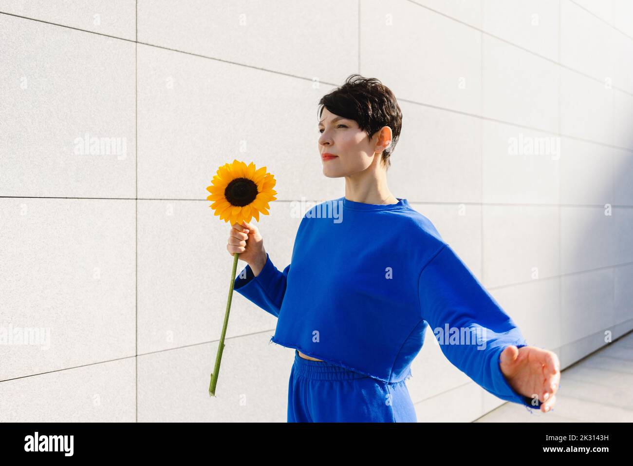 Frau mit kurzen Haaren hält Sonnenblume an der Wand an sonnigen Tag Stockfoto