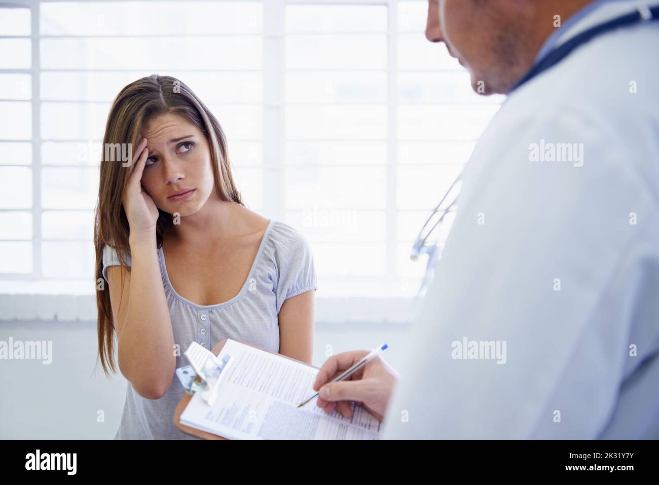 Also, wie lange haben Sie schon diese Kopfschmerzen. Eine junge Frau, die während eines Arztbesuches besorgt aussieht. Stockfoto