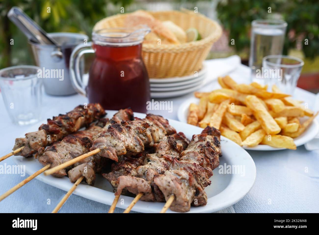 Selektiver Fokus auf Fleisch mit typischer griechischer Tischanordnung, einschließlich des halben Liter-Kännchens Rotwein, gebratener Kartoffeln, Souvlaki und Brot Stockfoto