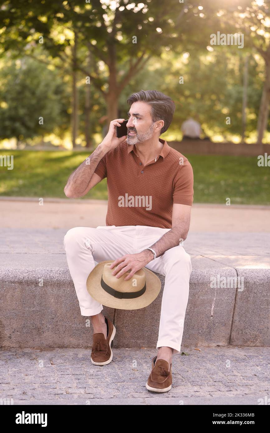 Entspannter Mann mit einem Hut in der Hand lächelnd, während er am Telefon im Freien in einem Park sitzt. Technologiekonzept. Stockfoto