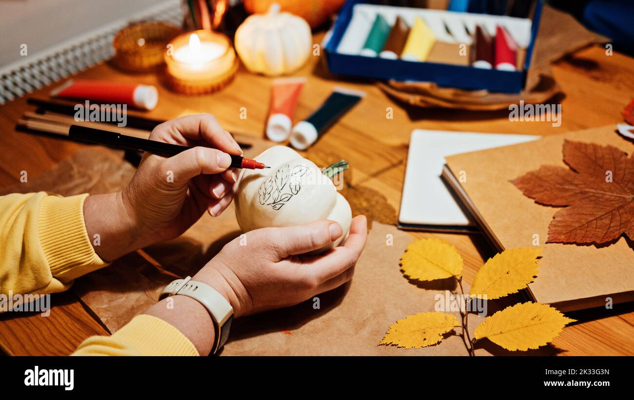 Herbstkürbisse Handwerk. DIY Herbst Crafts für Erwachsene. Weibliche Hände zeichnen Muster auf weißem Kürbis vor herbstlichem Dekorhintergrund auf den Tisch zu Hause Stockfoto