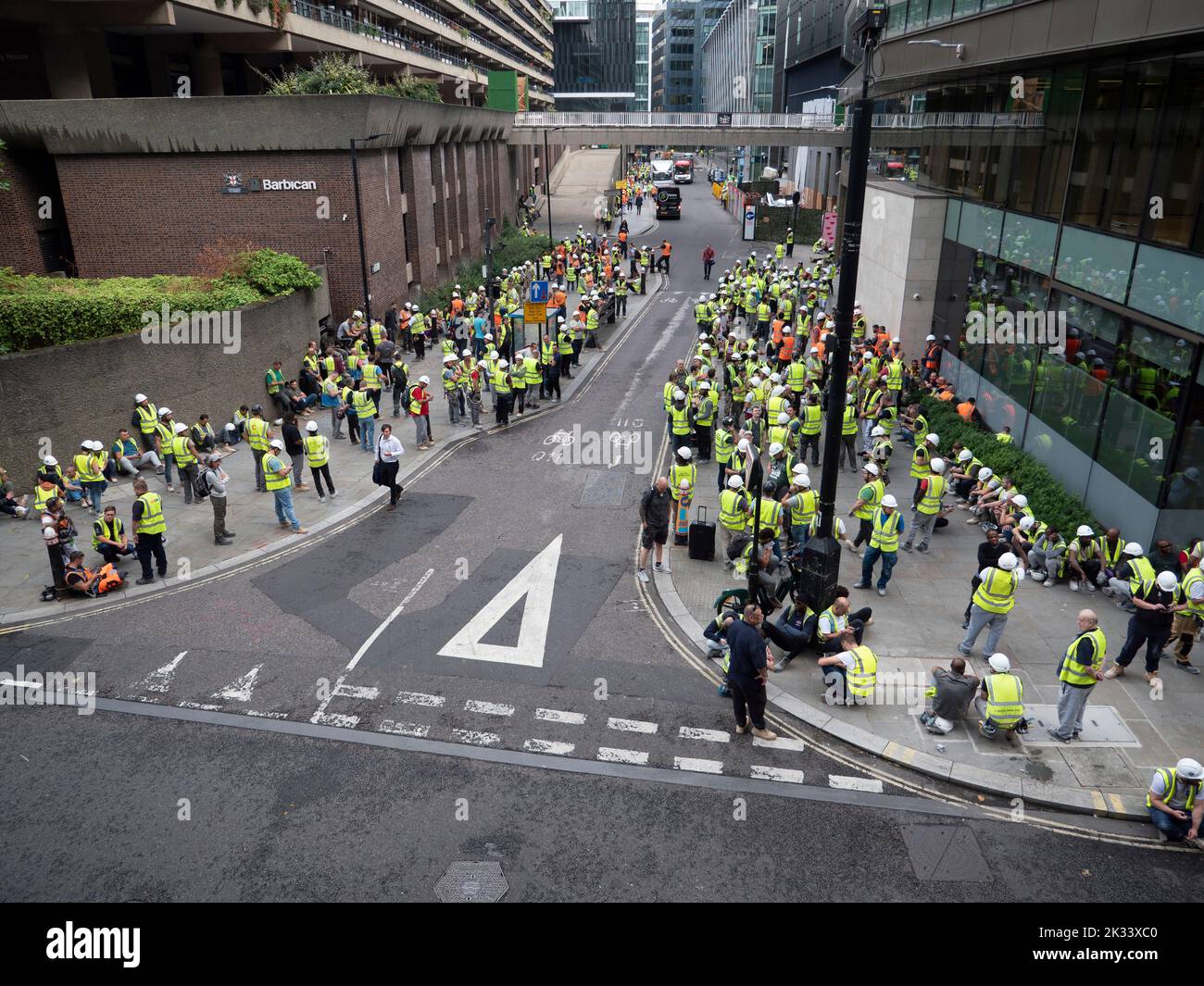 Massen von Bauarbeitern und Bauunternehmern, die Leuchtstoffjacken und Hüte tragen, versammeln sich während der Evakuierungsfeuerübung in Barbican London vor dem Gebäude Stockfoto
