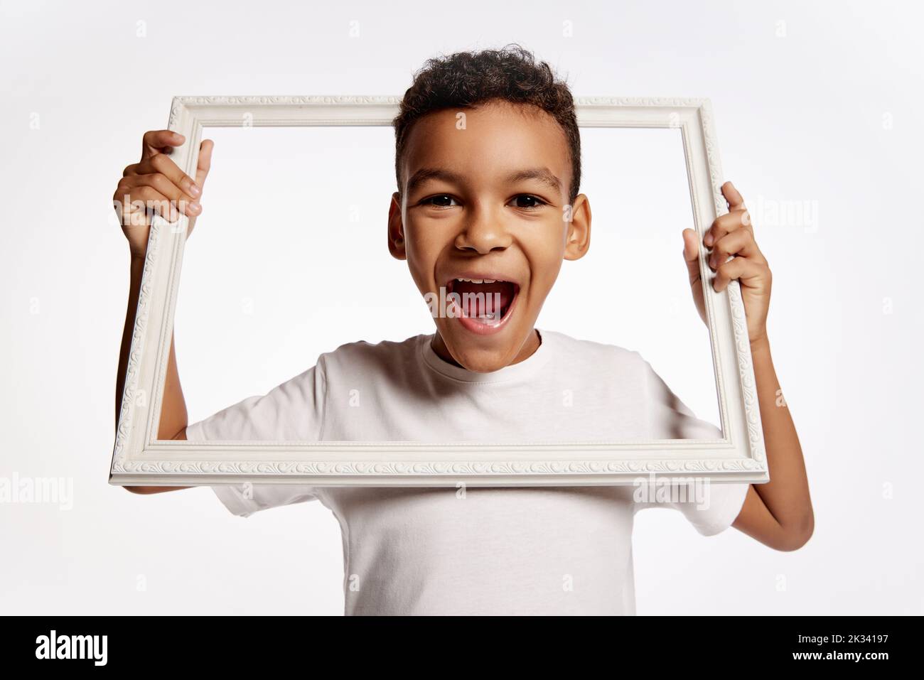 Fröhlicher glücklicher afrikanischer kleiner Junge in weißem T-Stück ragt aus dem Bilderrahmen und lacht isoliert auf weißem Hintergrund. Kinder Emotionen Konzept Stockfoto