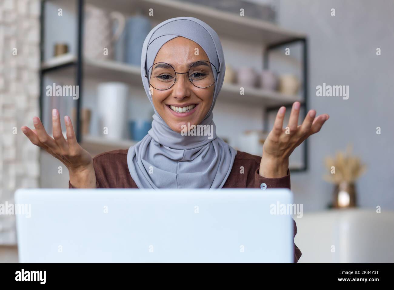 Videoanruf Online-Treffen mit Freunden, junge schöne muslimische Frau in Hijab, die Kamera anschaut und lächelt, mit Freunden aus der Ferne redet, Frau zu Hause in der Küche mit Laptop. Stockfoto