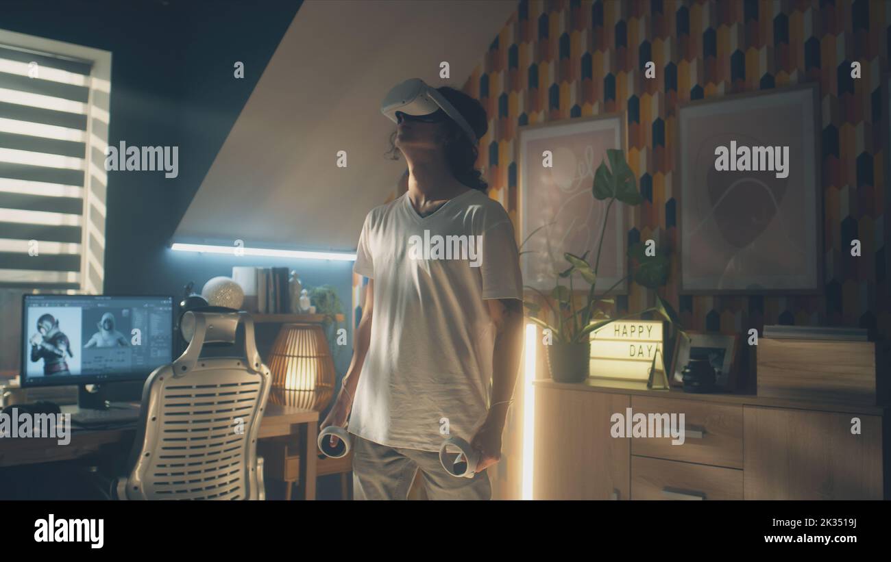 Mann, der in seiner Freizeit in seinem Zimmer Videospiele mit modernen VR-Headsets und drahtlosen Controllern spielt. Virtual-Reality-Gerät. Cyberspace und Metaverse Stockfoto