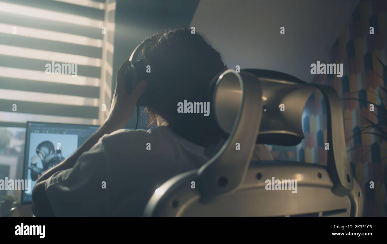 Rückansicht des Mannes in den Kopfhörern, der am Tisch sitzt und Videospiel-Helden und 3D-Animation in einem professionellen Computerprogramm erstellt, während er zuhause aus der Ferne arbeitet. Stockfoto