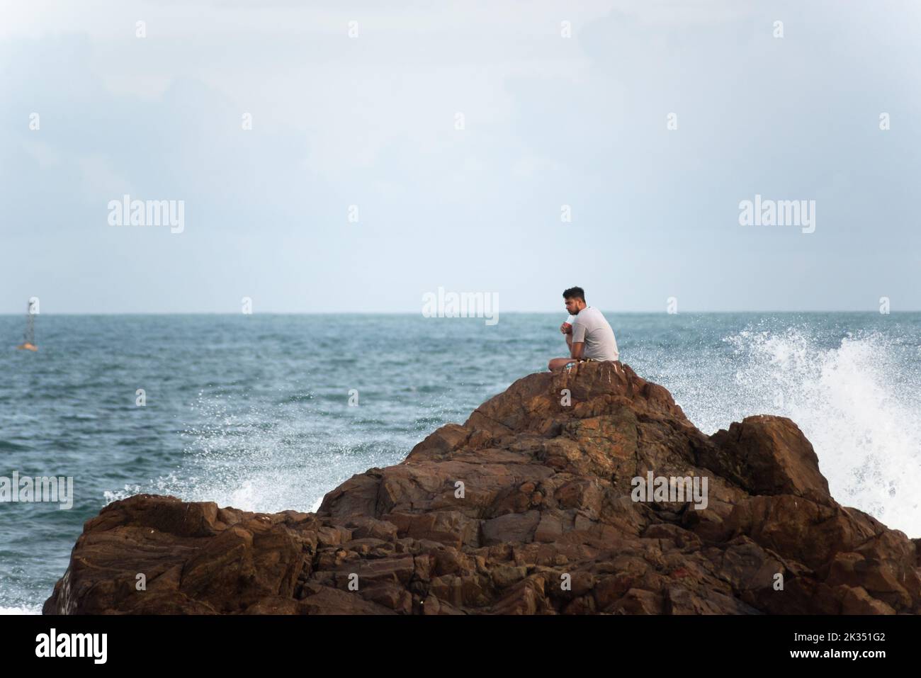 Ein junger Mann sitzt auf einem großen Felsen am Strand von Farol da Barra in Salvador, Bundesstaat Bahia, Brasilien Stockfoto