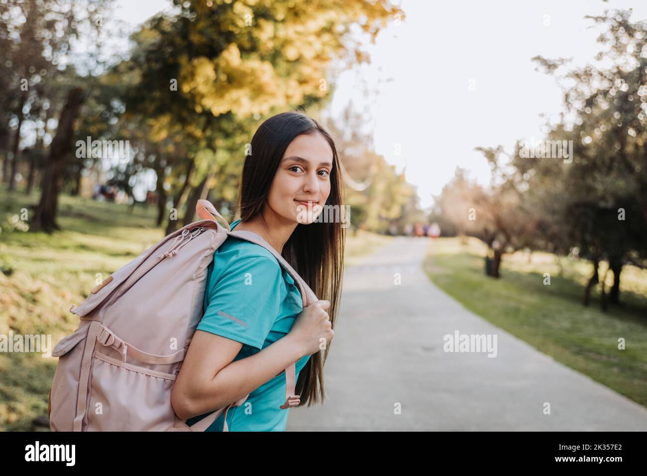 Rückansicht eines Studenten-Mädchens, das einen Rucksack trägt und mit einem ruhigen Lächeln in einer Parkstraße auf die Kamera blickt. Zukünftiger Weg Stockfoto