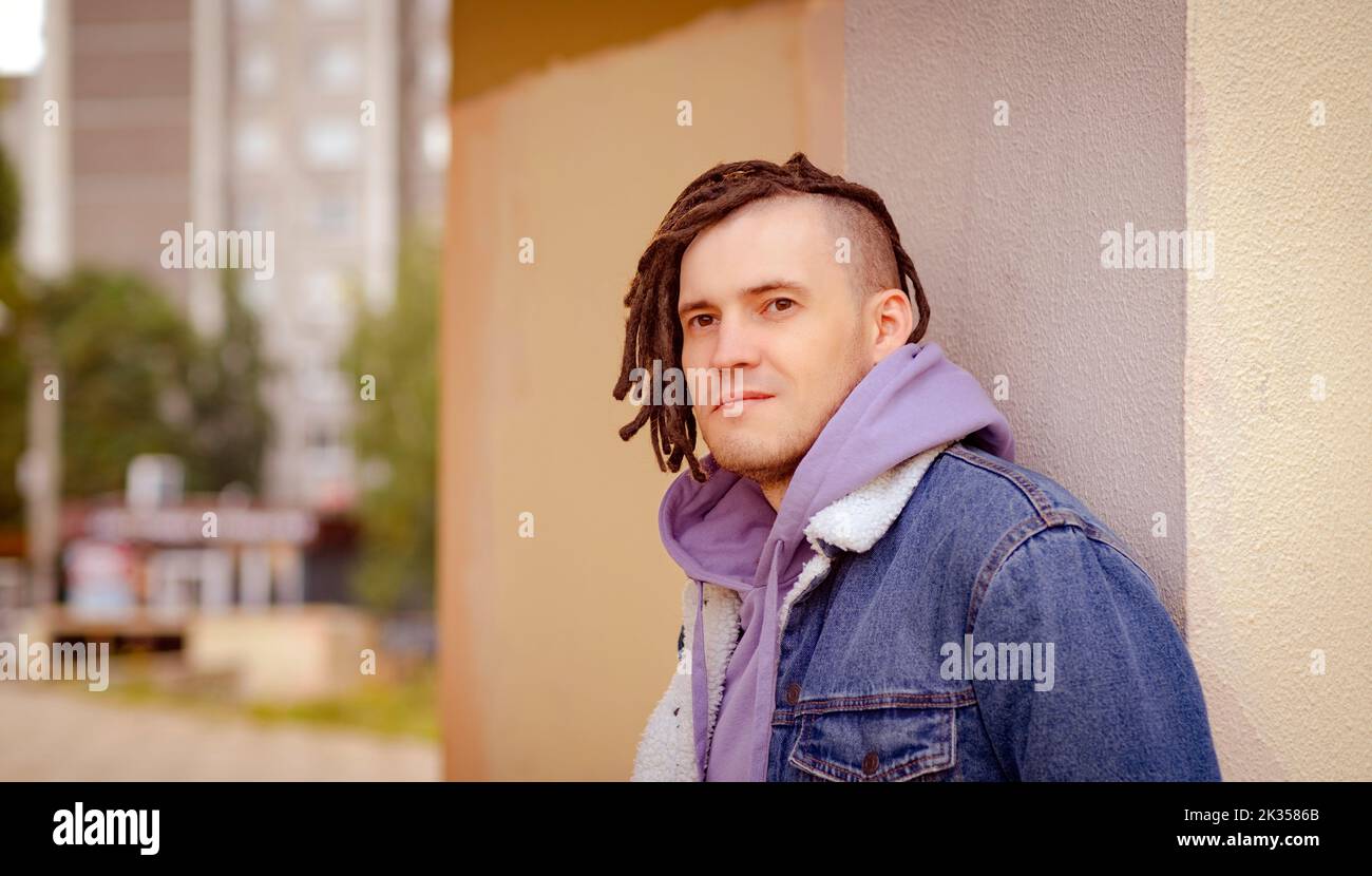 Porträt eines jungen, gutaussehenden Mannes mit Dreadlocks, die auf der Straße posieren, stehen und sich an einer Wand lehnen Stockfoto