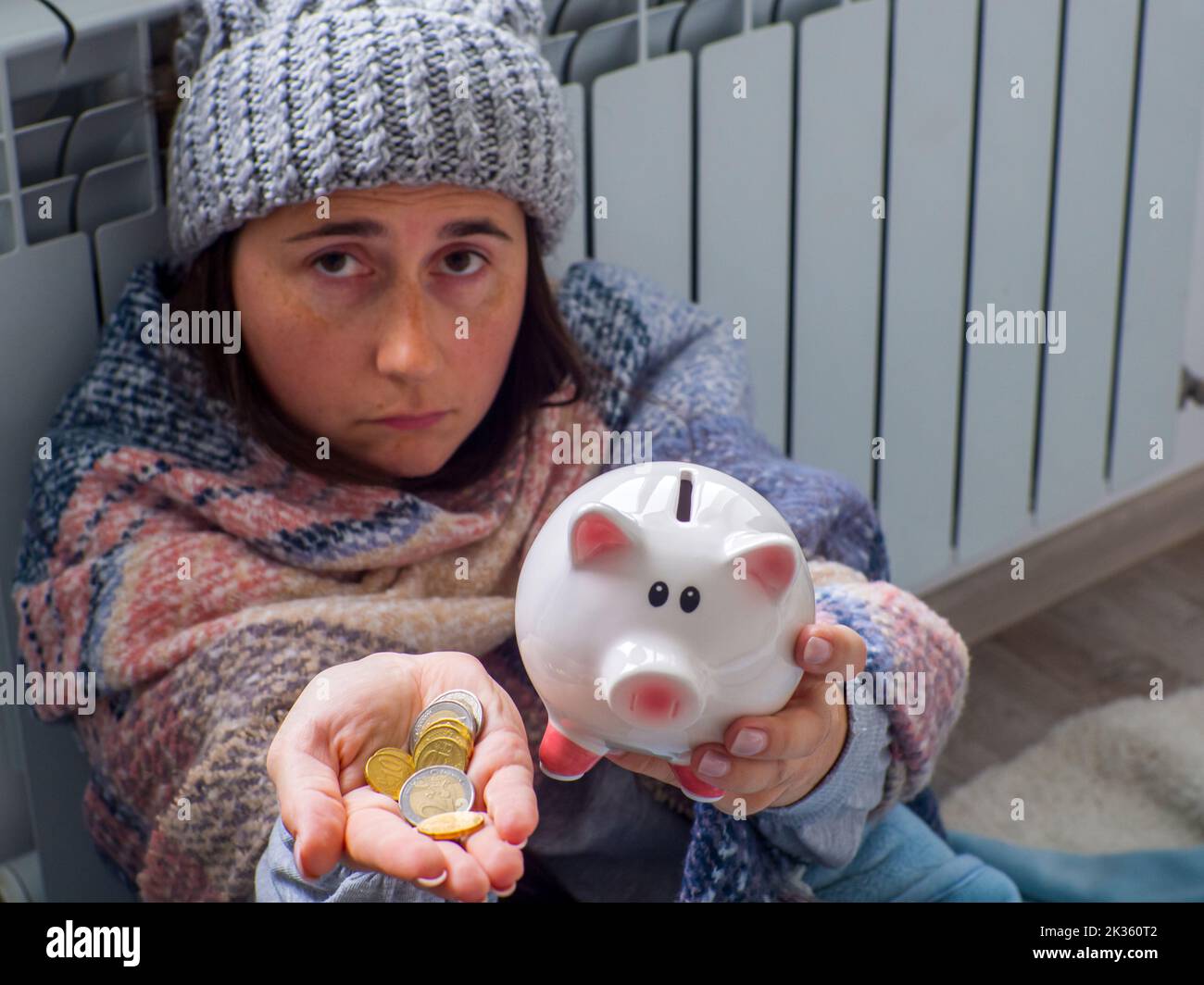 Frau zählt Euromünzen und hält Sparschwein in der Nähe der Batterie zu Hause. Warm gekleidete Frau ist über den hohen Preis der Heizung aufgeregt. Teuer en Stockfoto