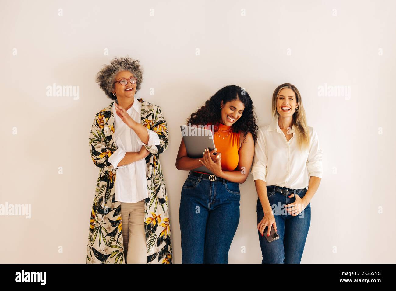 Erfolgreiche Geschäftsfrauen lachen fröhlich, während sie vor einem Wandhintergrund stehen. Eine Gruppe von drei multikulturellen Geschäftsfrauen, die in einem zusammenarbeiten Stockfoto