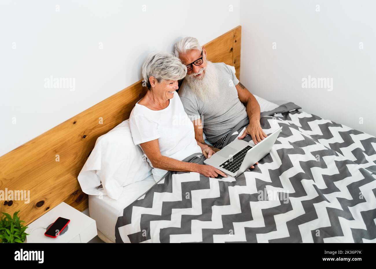 Glückliches Seniorenpaar, das Spaß mit dem Computer hat, während es unter den Decken im Bett lag - Technologie und Lifestyle-Konzept für ältere Menschen Stockfoto