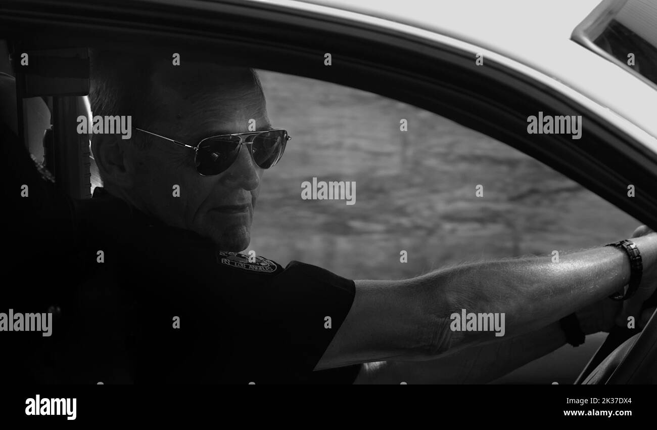 Performer, der als amerikanischer Polizeibeamter in Los Angeles in Spiegelsbrillen gekleidet ist und während des Christchurch Festivals in Großbritannien ein Polizeifahrzeug fährt Stockfoto