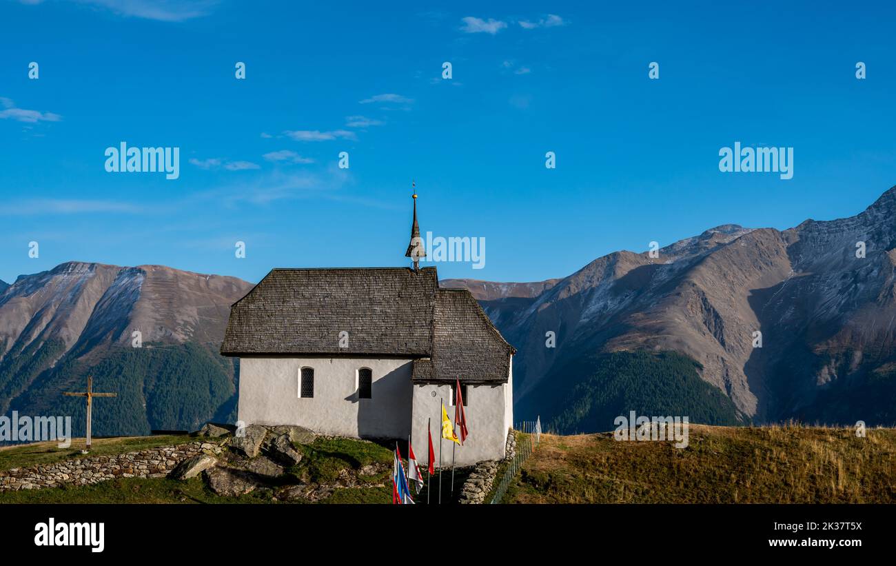 Landschaft aus Kirche und Bergen. Schweizer Bettmeralp-Kirche mit Schweizer Kantonsflaggen. Ruhige Szene. Stockfoto