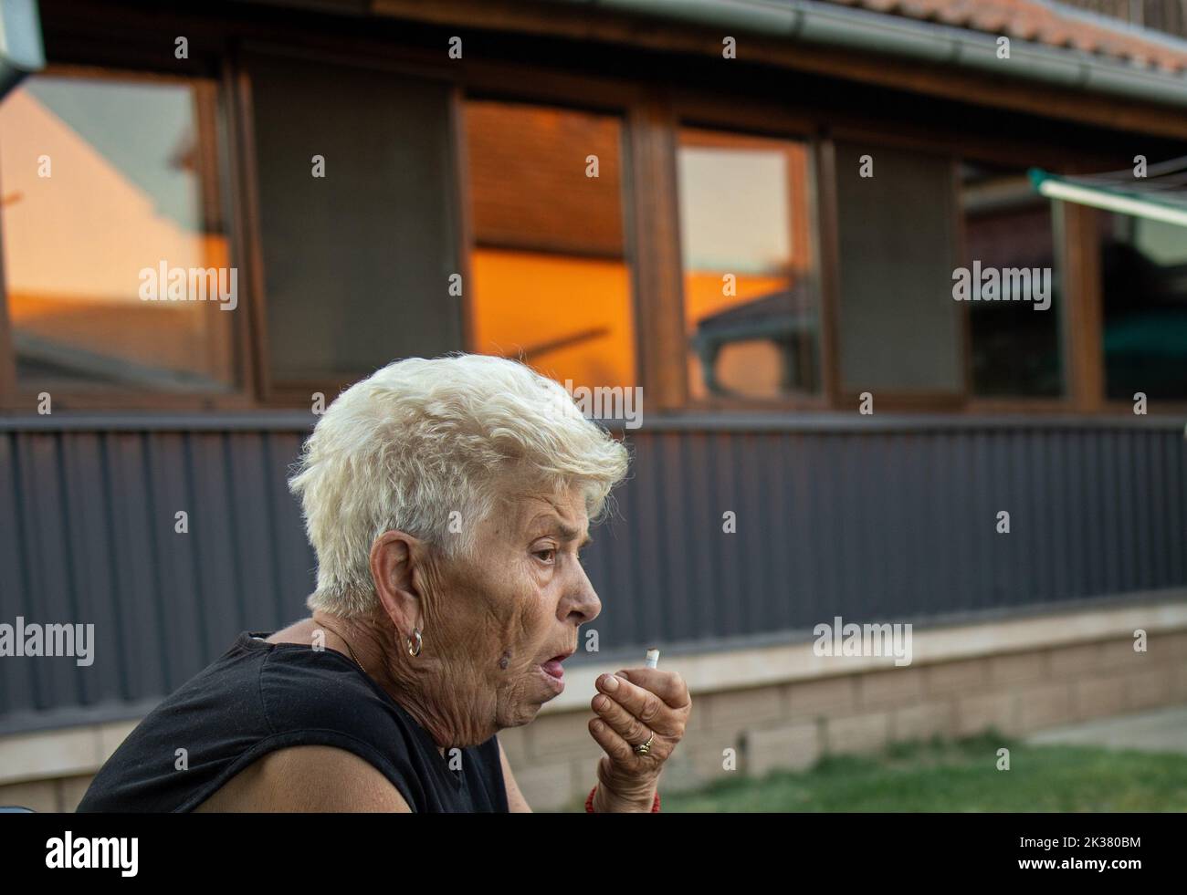 Eine ältere Frau sitzt auf einem Stuhl und hustet und hält eine Zigarette in der Hand. Gesundheitsfürsorge, Warnung für Raucher Stockfoto