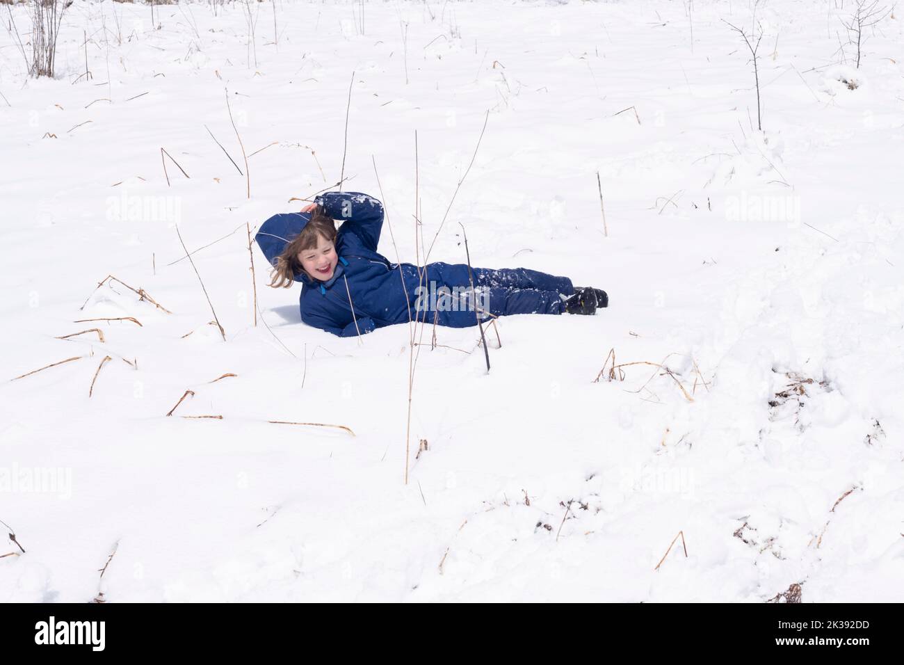 Ein lachendes Mädchen in einem blauen Schneeanzug rollt einen Hügel mit frisch gefallener Schneedecke im Süden Ontarios hinunter. Stockfoto