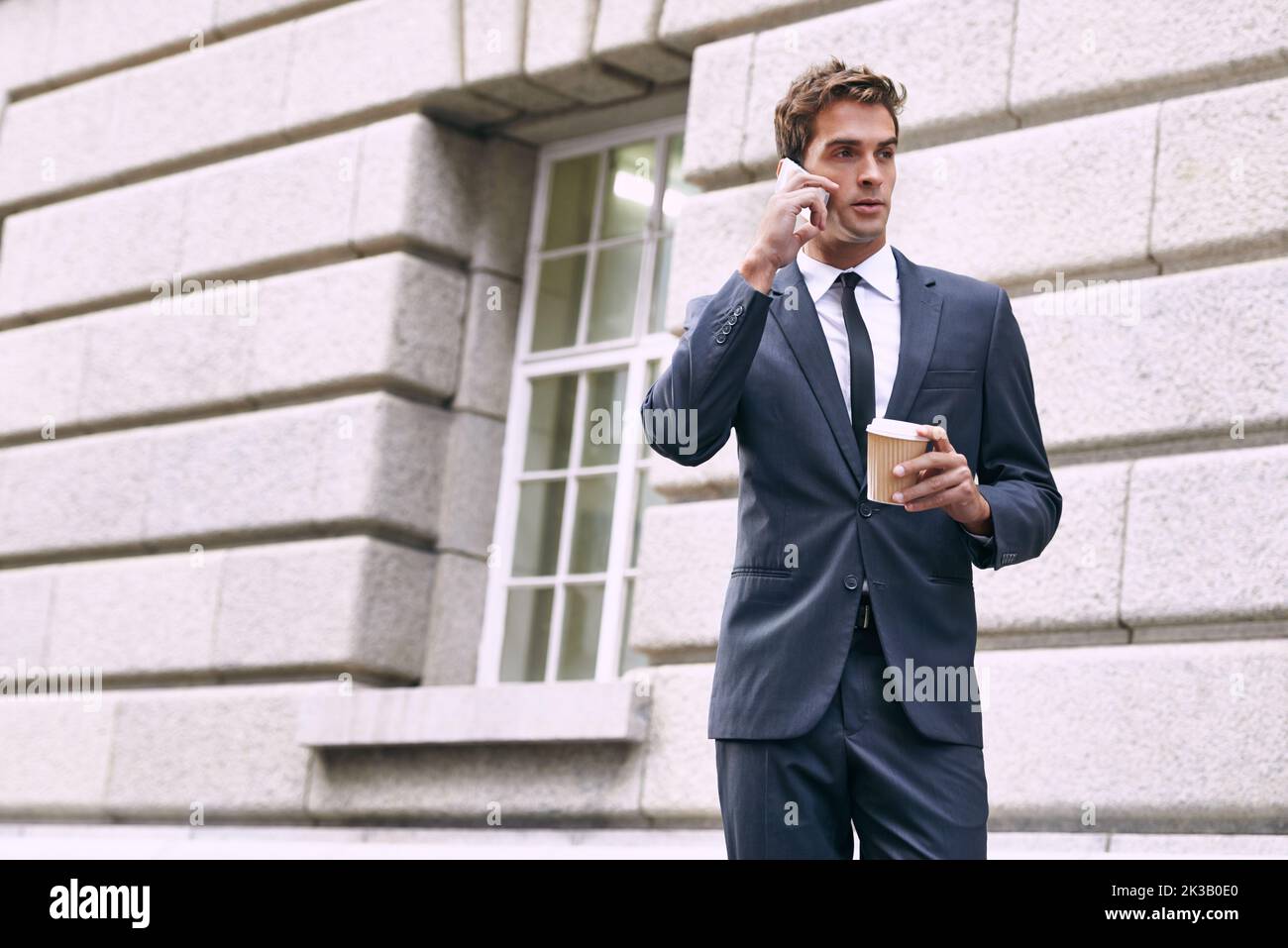 Unterwegs. Ein gutaussehender Geschäftsmann, der mit einem Kaffee zum Mitnehmen in der Hand telefoniert. Stockfoto