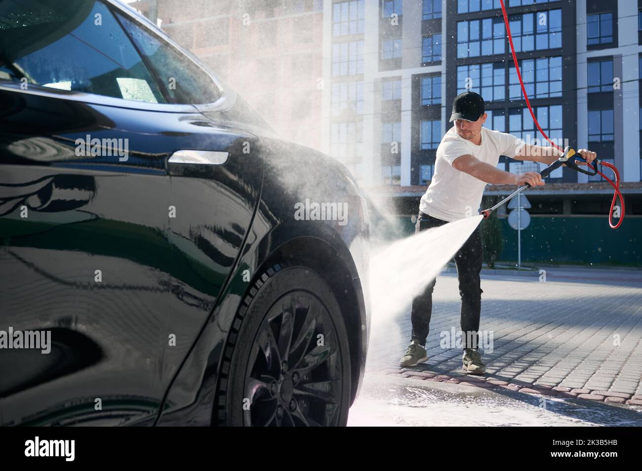 Guy in Cap Reinigung seines schwarzen Autos mit Hochdruckwasser im Service. Waschen von Auto mit fließendem Wasser auf dem Hintergrund des Wohngebäudes. Stockfoto