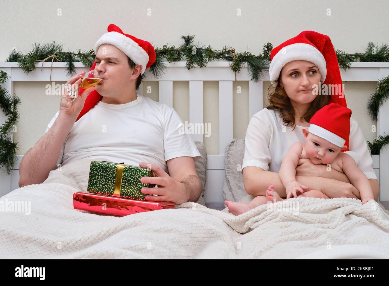 Ein Mann trinkt Alkohol, während eine Frau mit einem Kind in einem Bett sitzt, das zu Weihnachten und Neujahr dekoriert ist, ein Familienpaar in einem Schlafzimmer Stockfoto