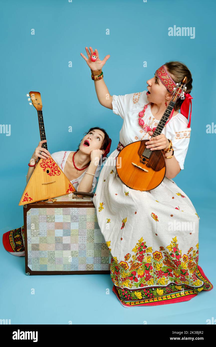 Musikerinnen in russischen Volkskleidern mit Musikinstrumenten auf blauem Studiohintergrund. Glückliche Künstler aus Russland in weißer Nationalkleidung mit Stockfoto
