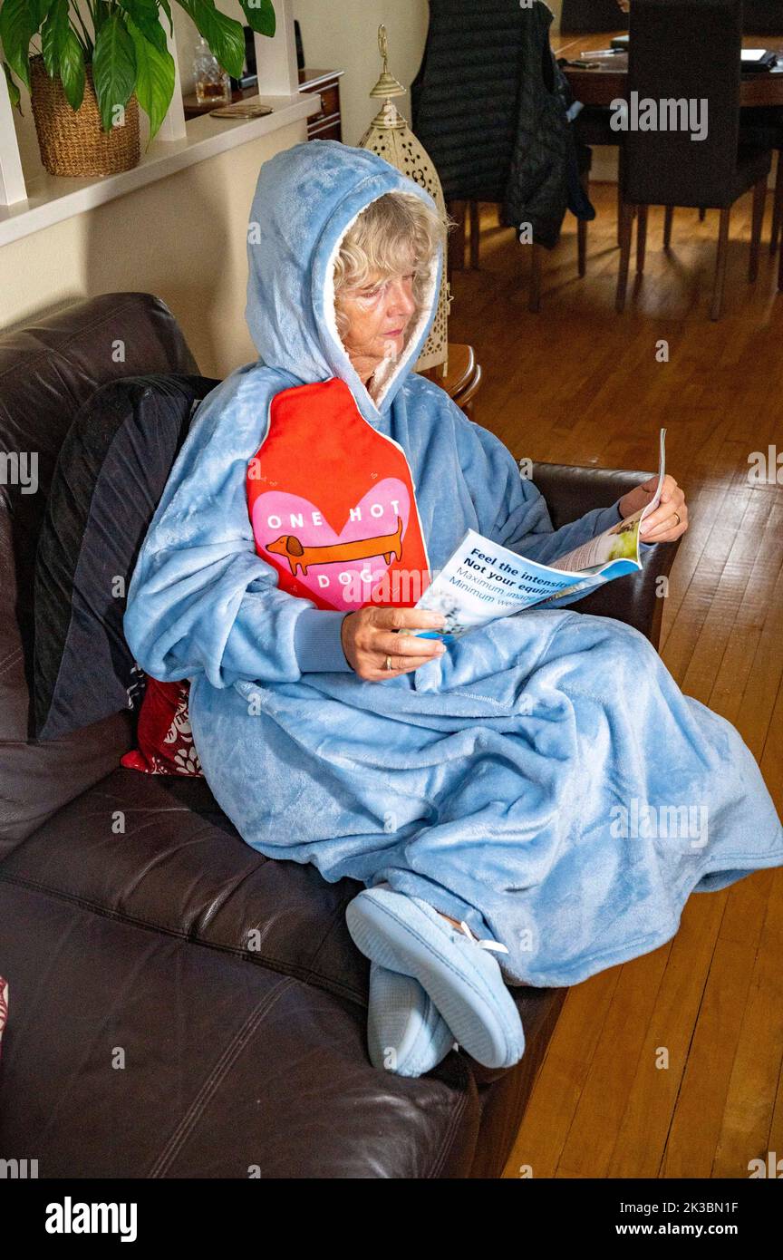 Frau trägt ein Fleece mit Kapuze oder Hoodie und hält eine Wärmflasche, um sich zu Hause warm zu halten, wenn die Energiekosten in Großbritannien steigen (modellhaft) Stockfoto